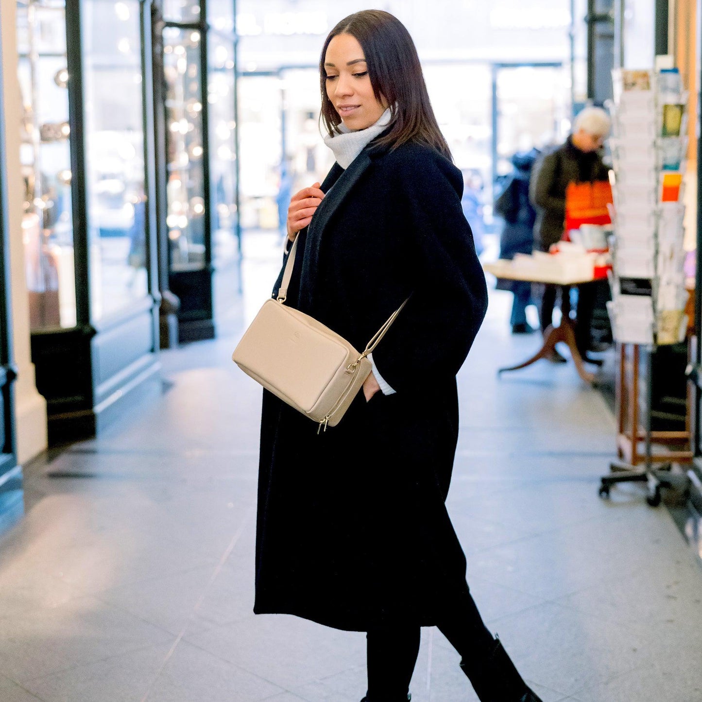 Eine junge Frau trägt in einer Einkaufspassage eine Crossbody in sand über ihrem dunklen Mantel