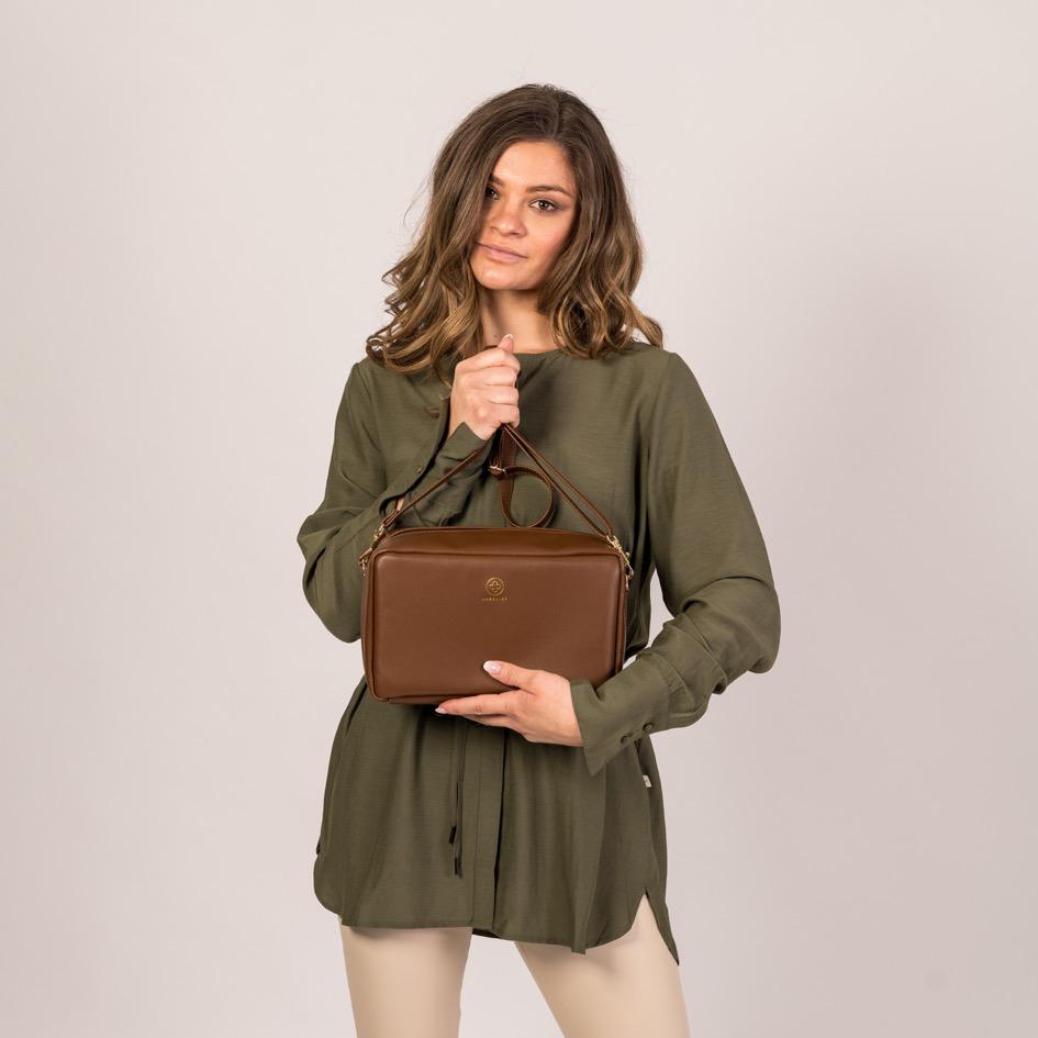 Eine junge Frau hält sich die Crossbody ZOE XL in coffee vor den Bauch und hält mit einer Hand den Tragegurt hoch