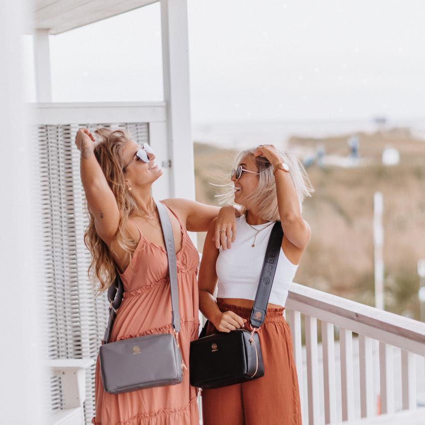 Zwei junge Frauen lachen sich an und zeigen die Crossbody in graphite und in schwarz