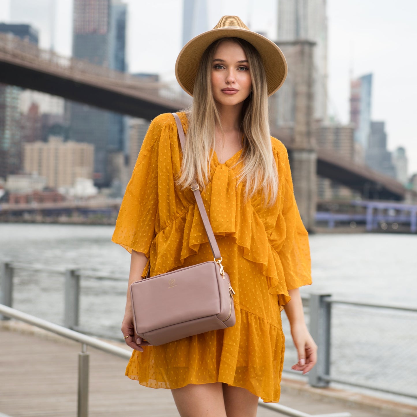 Die Crossbody ZOE XL in puder wird von einer Frau in strahlend gelben Kleid getragen, sie trägt einen Hut und im Hintergrund ist die Brooklyn Bridge zu sehen.