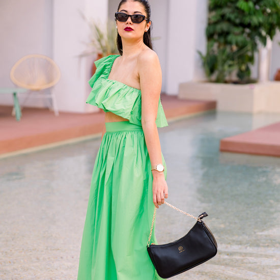 Ein Model trägt einen grünen Zweiteiler mit Midi-Rock und steht mit dem Füße im Pool, an der Hand die Handtasche an der Kette.