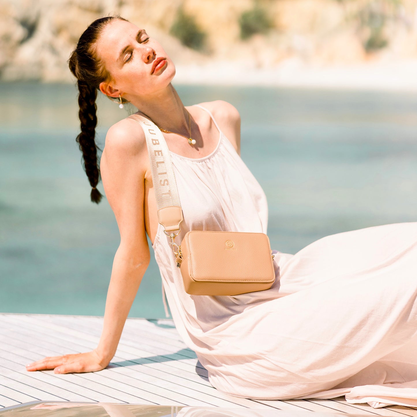 Frau sitzt mit einem simplen weißen Kleid vorm Hintergrund mit Meer, sie trägt den Signatur Strap an der Jade