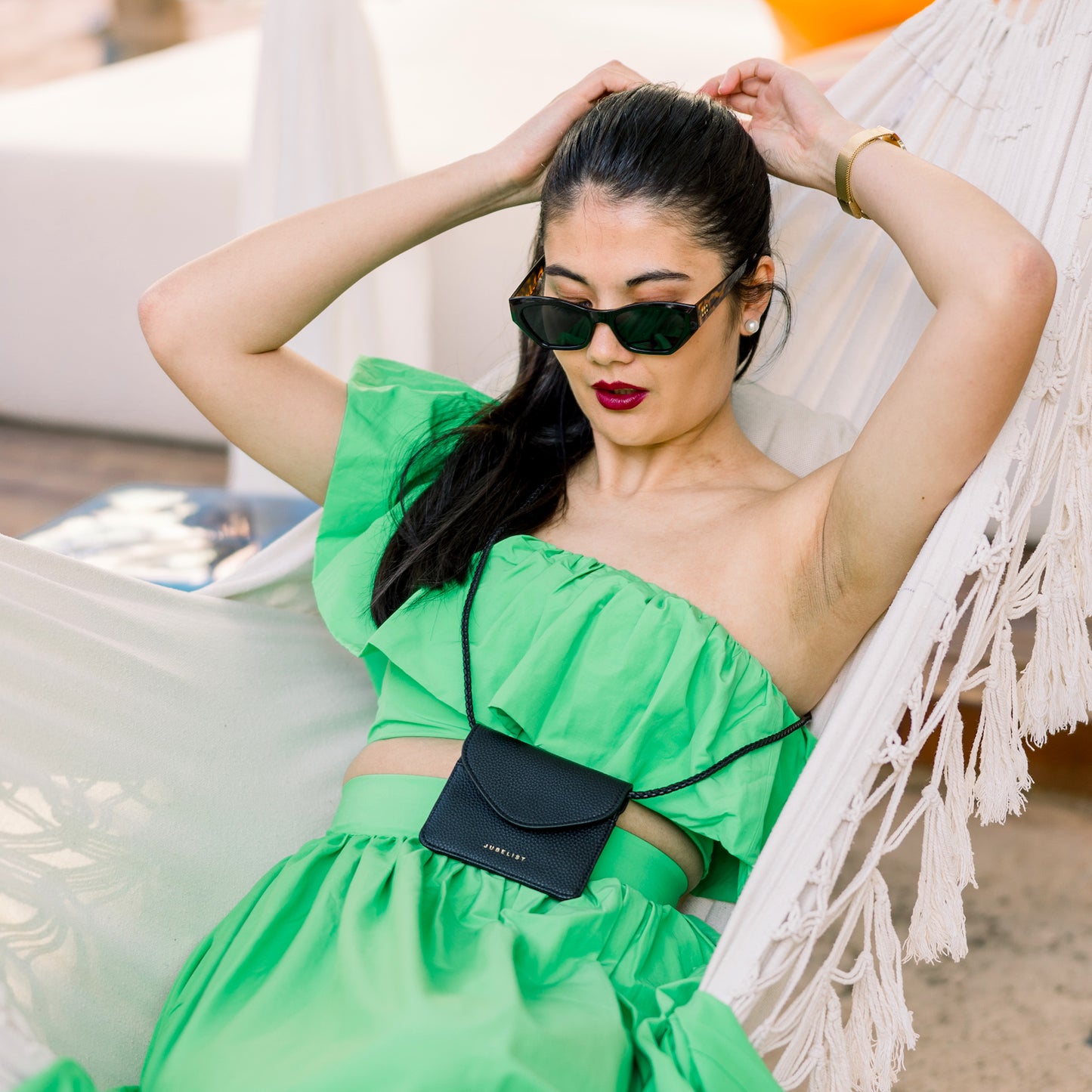 Ein Model liegt in einem grünen extravaganten grünen Kleid auf der Hängematte und zeiht ihrem Zopf stramm. Sie trägt BAILY crossbody in schwarz.