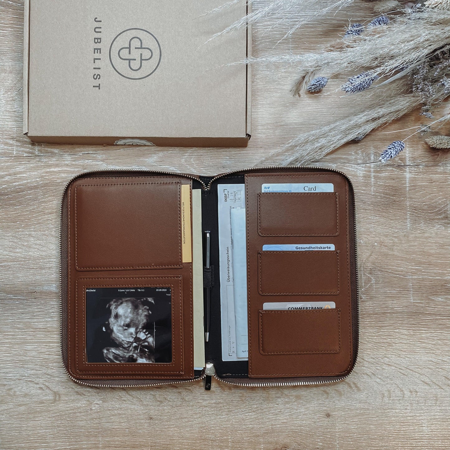 Eine Dokumententasche in coffee liegt aufgeklappt auf einem Tisch und ist gefüllt mit Krankenkassen- und EC Karte, mit einem Stift, mit einem Ultraschallbild im Sichtfenster , Impfpass und Mutterpass finden ebenfalls Platz