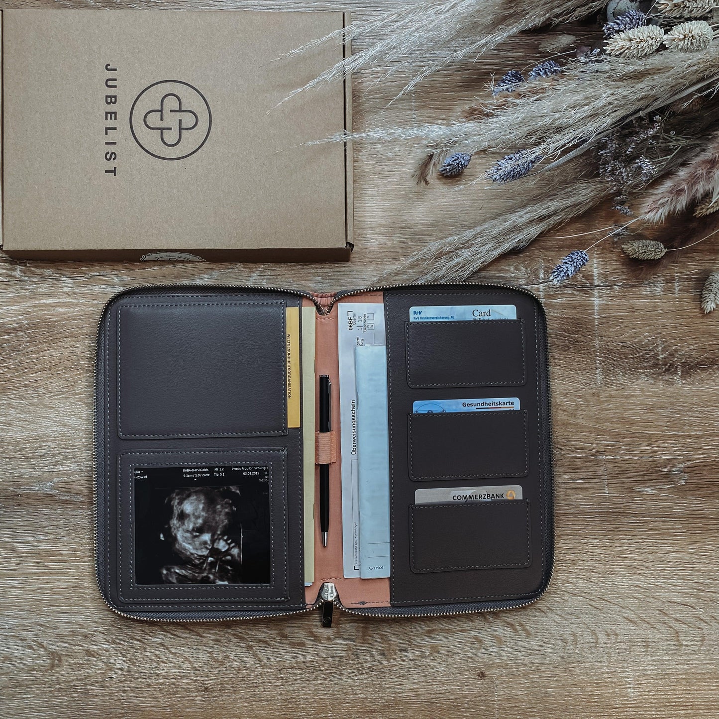 Eine Dokumententasche in graphite mit rosa Innenfutter liegt aufgeklappt auf einem Tisch und ist gefüllt mit Krankenkassen- und EC Karte, mit einem Stift, mit einem Ultraschallbild im Sichtfenster , Impfpass und Mutterpass finden ebenfalls Platz