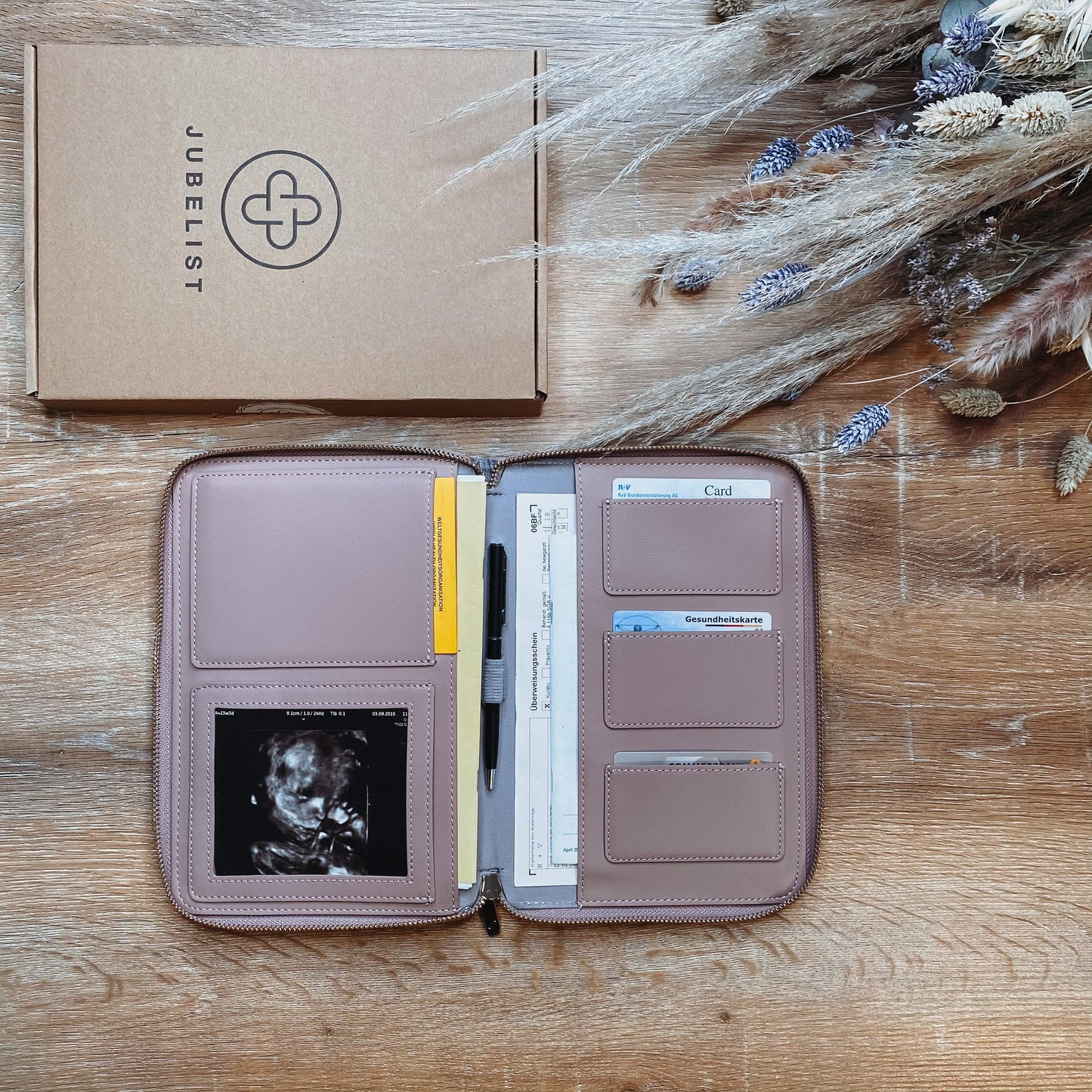 Eine Dokumententasche in puder liegt aufgeklappt auf einem Tisch und ist gefüllt mit Krankenkassen- und EC Karte, mit einem Stift, mit einem Ultraschallbild im Sichtfenster , Impfpass und Mutterpass finden ebenfalls Platz