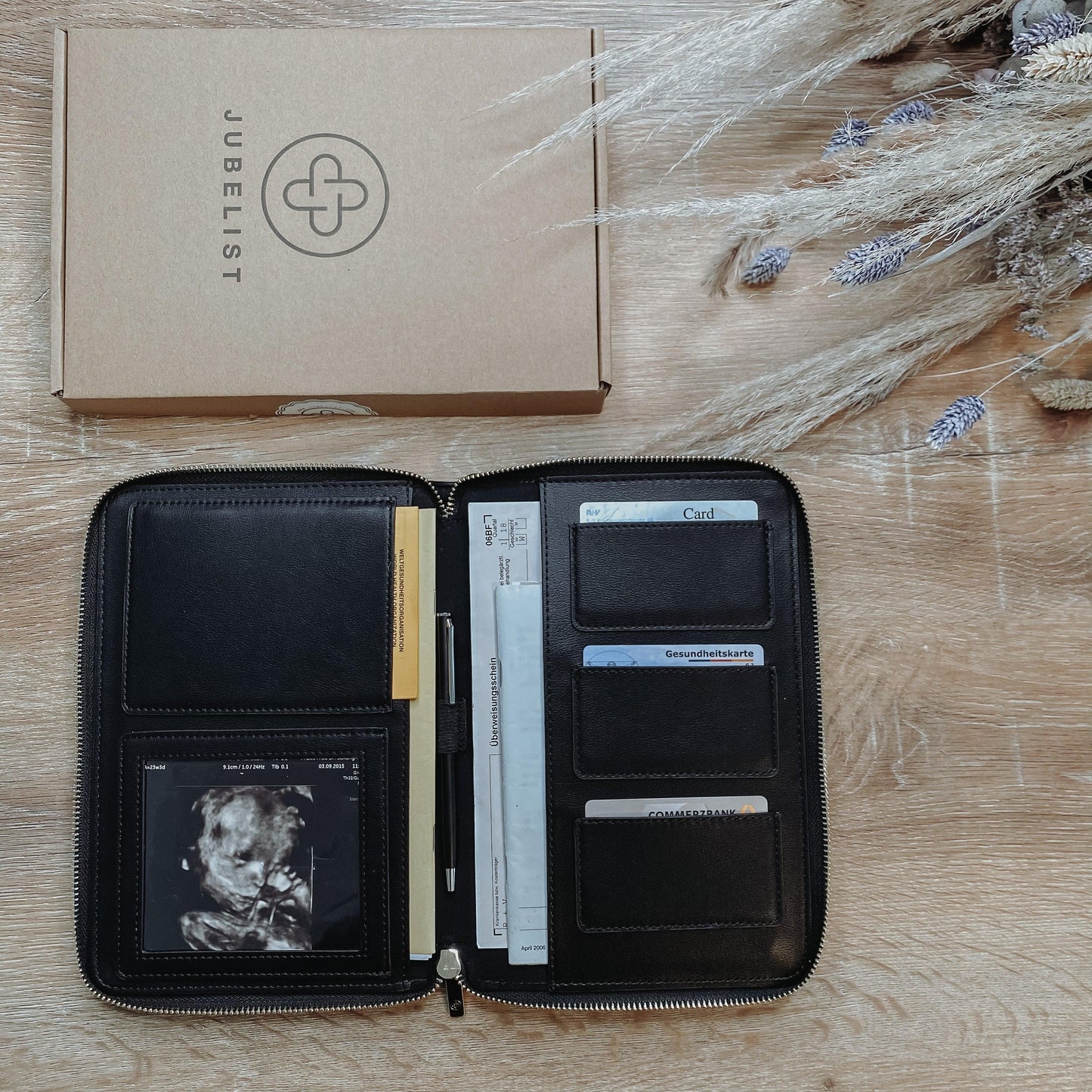 Eine Dokumententasche in schwarz silber liegt aufgeklappt auf einem Tisch und ist gefüllt mit Krankenkassen- und EC Karte, mit einem Ultraschallbild im Sichtfenster , Impfpass und Mutterpass finden ebenfalls Platz