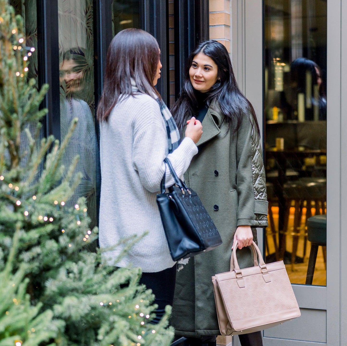 Zwei junge Frauen unterhalten sich und tragen beide die Business Bag Logo Edition in schwarz und taupe am Griff und über den Unterarm