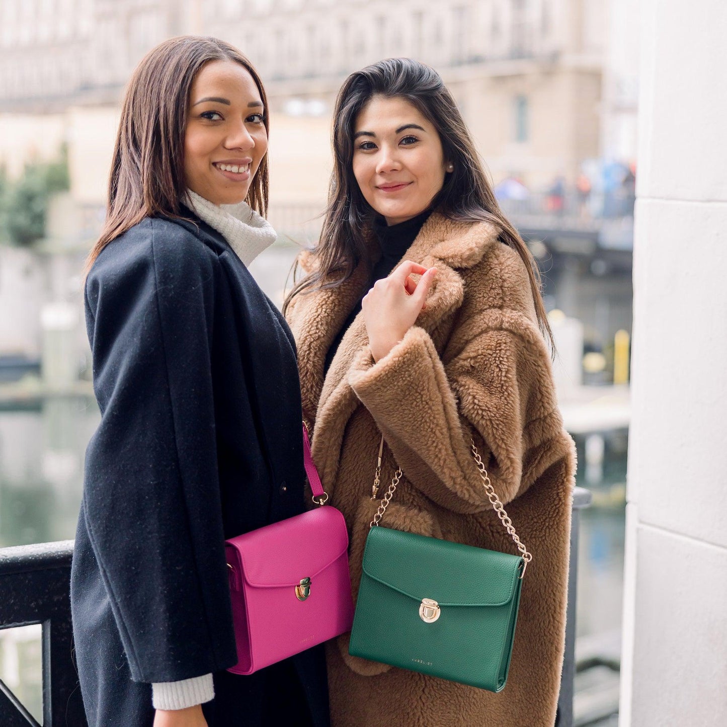 Zwei junge Frauen stehen nah beieinander und eine trägt ihre Tasche in hot pink crossbody und die andere in grün mit der Kette über den Unterarm