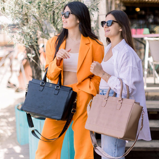 Zwei Frauen schauen gemeinsam in eine  Richtung, die Frau mit der Business Bag Logo Edition in schwarz trägt einen orangenen Hosenanzug und die Frau mit der taupe farbenen Business Bag Logo Edition trägt eine hellblaue Jeans und eine weiße Bluse