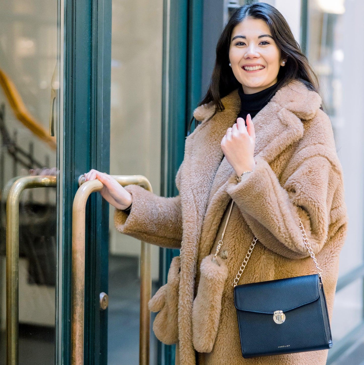 Eine junge Frau trägt ihre Tasche in schwarz über ihrem Unterarm und strahlt in die Kamera