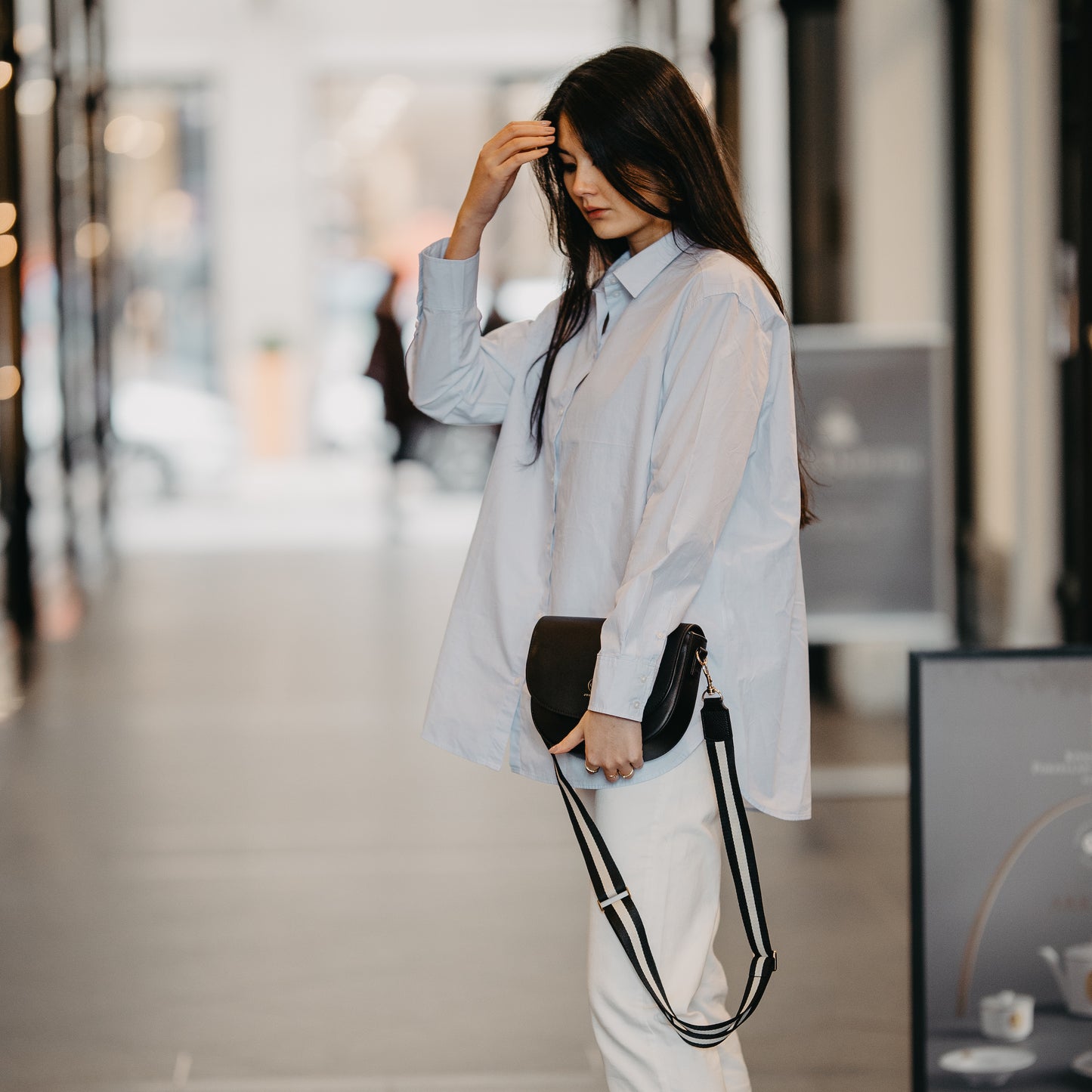 In einer Einkaufspassage steht eine junge Frau seitlich und hält die Saddle Bag in der Hand, der Taschengurt  Universal Strap in schwarz-weiß hängt locker runter