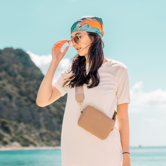 Ein Model mit dunklen Haaren trägt Sonnenbrille und ein buntes Kopftuch, sie trägt die Mini Crossbody JADE in taupe.