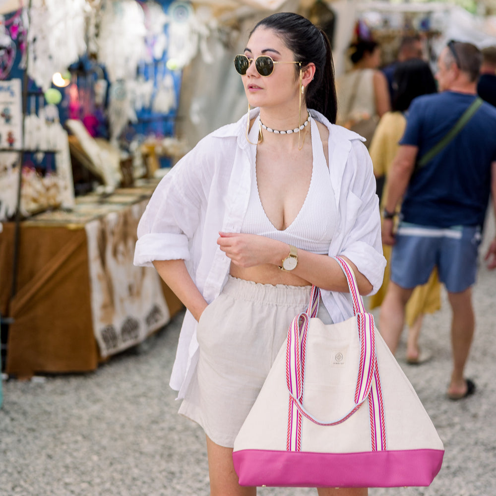 
                  
                    Ein Model mit beigen Shorts und weißem Hemd hat die Canvas Bag mit Pinke Boden und gestreiften Henkeln über den Arm.
                  
                