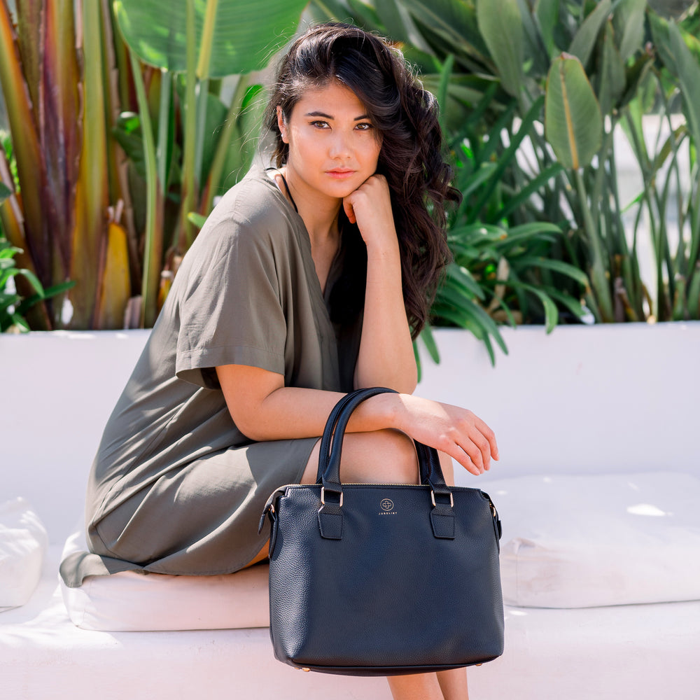 Ein Model sitzt auf einer weißen Bank, im Hintergrund grüne Blätter. Mit einem schlichten dunkelgrünen Kleid trägt sie die Handtasche Grace in schwarz mit goldenen Details.