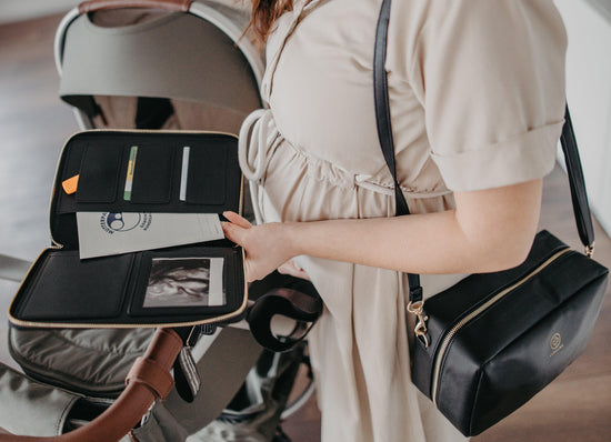 Schwangere Frau hält Dokumententasche mit Mutterpass, trägt eine Wickeltasche ZOE. Im Hintergrund ist ein Kinderwagen zu sehen.