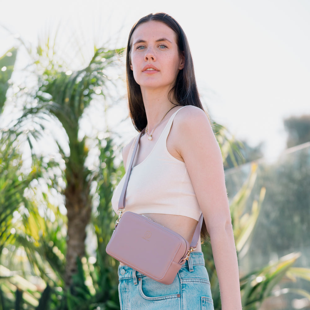 
                  
                    Ein Model mit dunklen Haaren schaut in die Kamera, trägt die Mini Crossbody JADE in puder. Sie hat blaue Jeansshorts und ein weißes Top.
                  
                