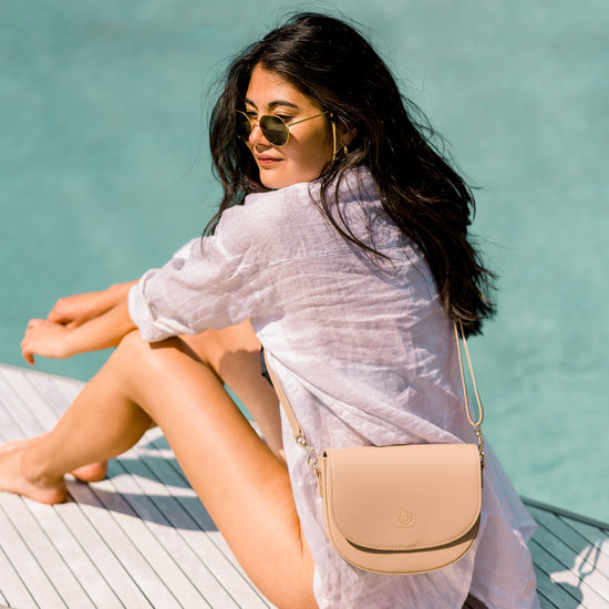 Model sitzt an einem Steg, im Hintergrund das Wasser. Mit offenen Haaren, weißem Leinenhemd und einer Sonnenbrille, Die sandfarbene Saddle Bag trägt sie crossbody.