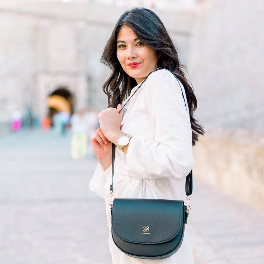 
                  
                    Das Model mit dunklen Haar und einem weißen Kleid strahlt in die Kamera und trägt dabei die Saddle Bag in schwarz.
                  
                