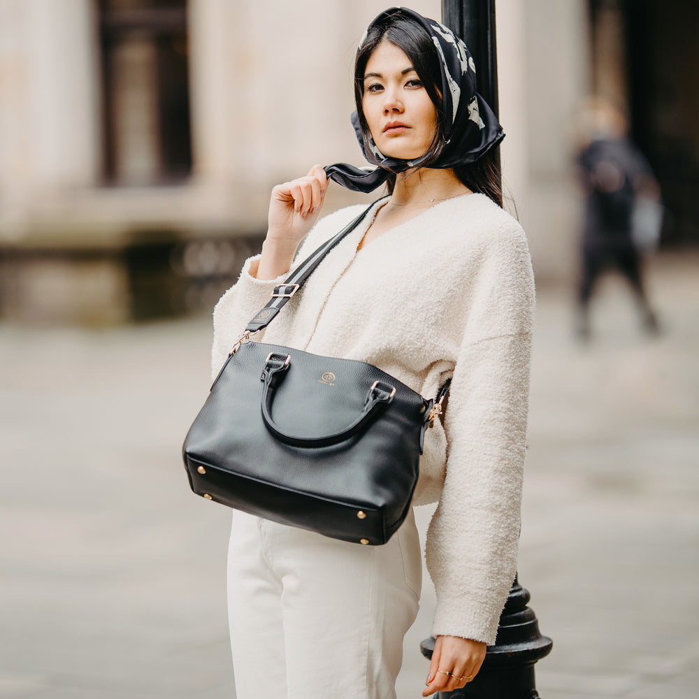 
                  
                    Eine junge Frau trägt ihre Grace in schwarz crossbody, sie lehnt gegen eine Laterne und hat ein schwarzes Tuch um die Haare gebunden
                  
                
