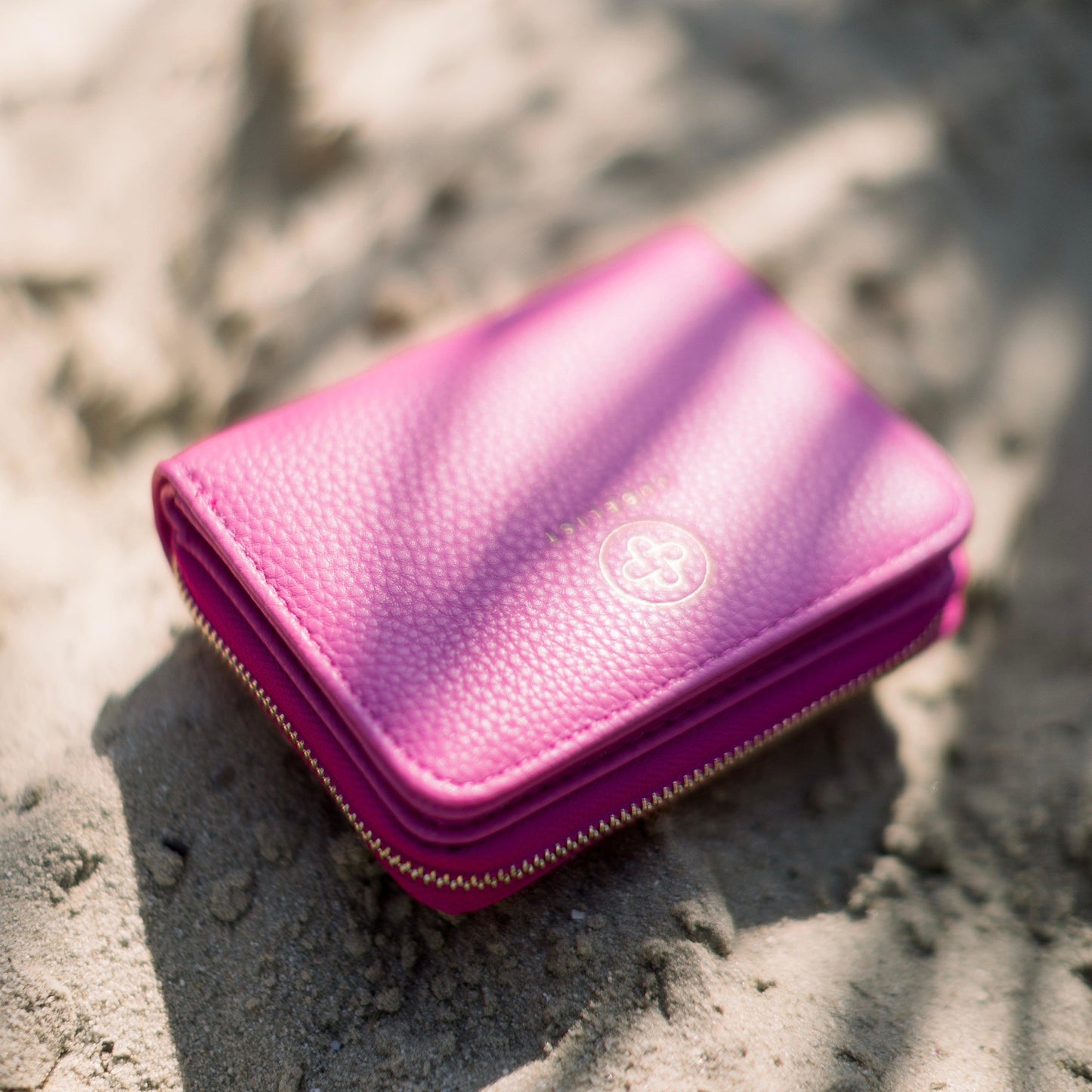 das Portemonnaie in hot pink liegt im Sand und die Sonne wirft Schattenmuster 