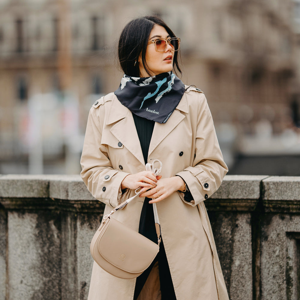 
                  
                    Eine Frau steht und hält in einer Hand die Saddle Bag am Universal Strap in beige weiß fest
                  
                