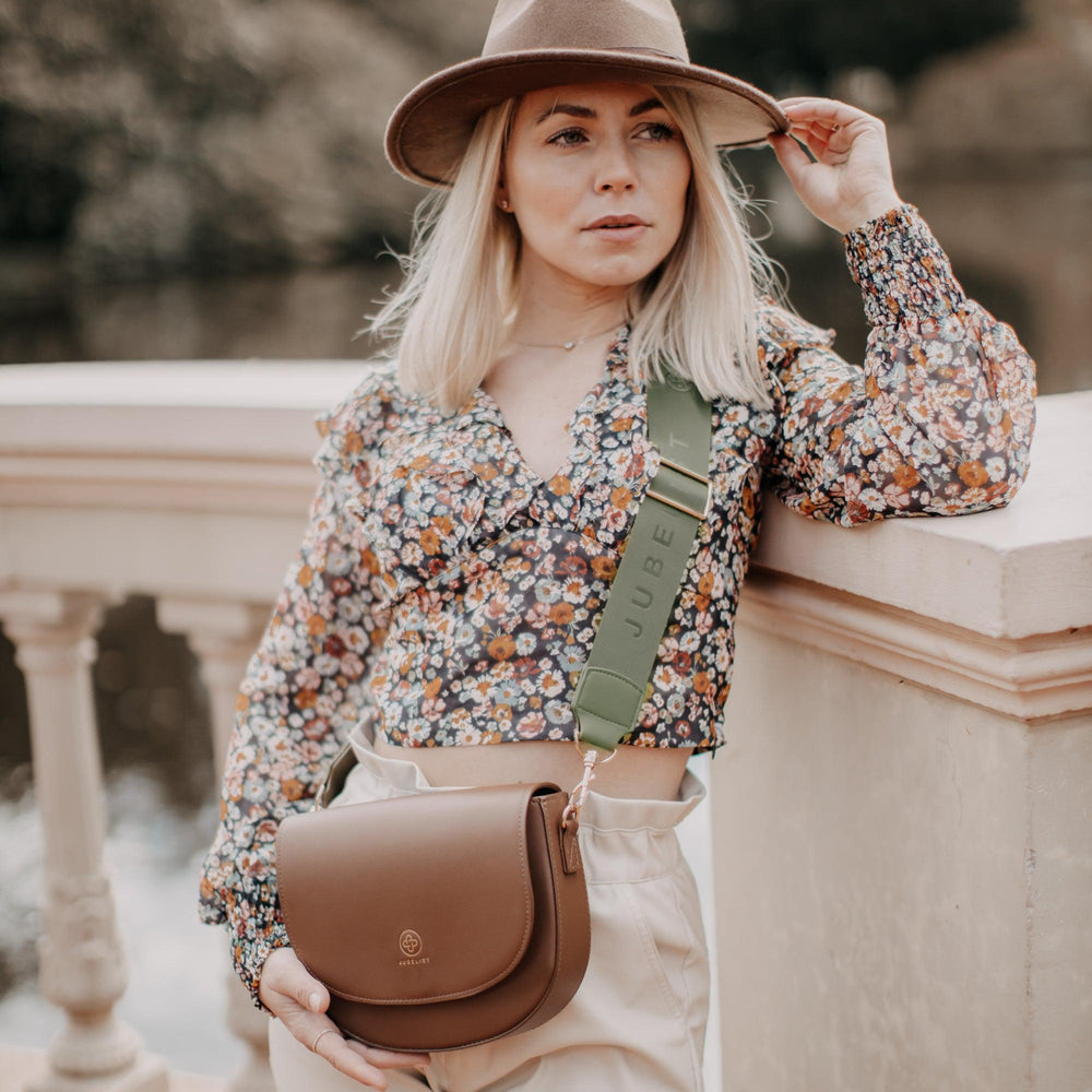 
                  
                    Eine blonde Frau lehnt an einer Mauer und trägt eine Saddle Bag in Coffee crossbody mit einem Fashion Strap in khaki
                  
                