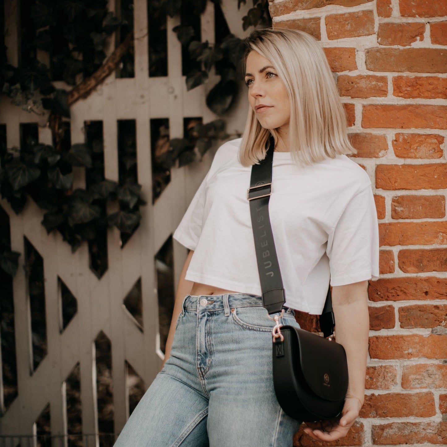 Das blonde Model trägt Jeans und T-Shirt kombiniert mit einer schwarzen Saddle Bag und einem Fashion Strap in schwarz