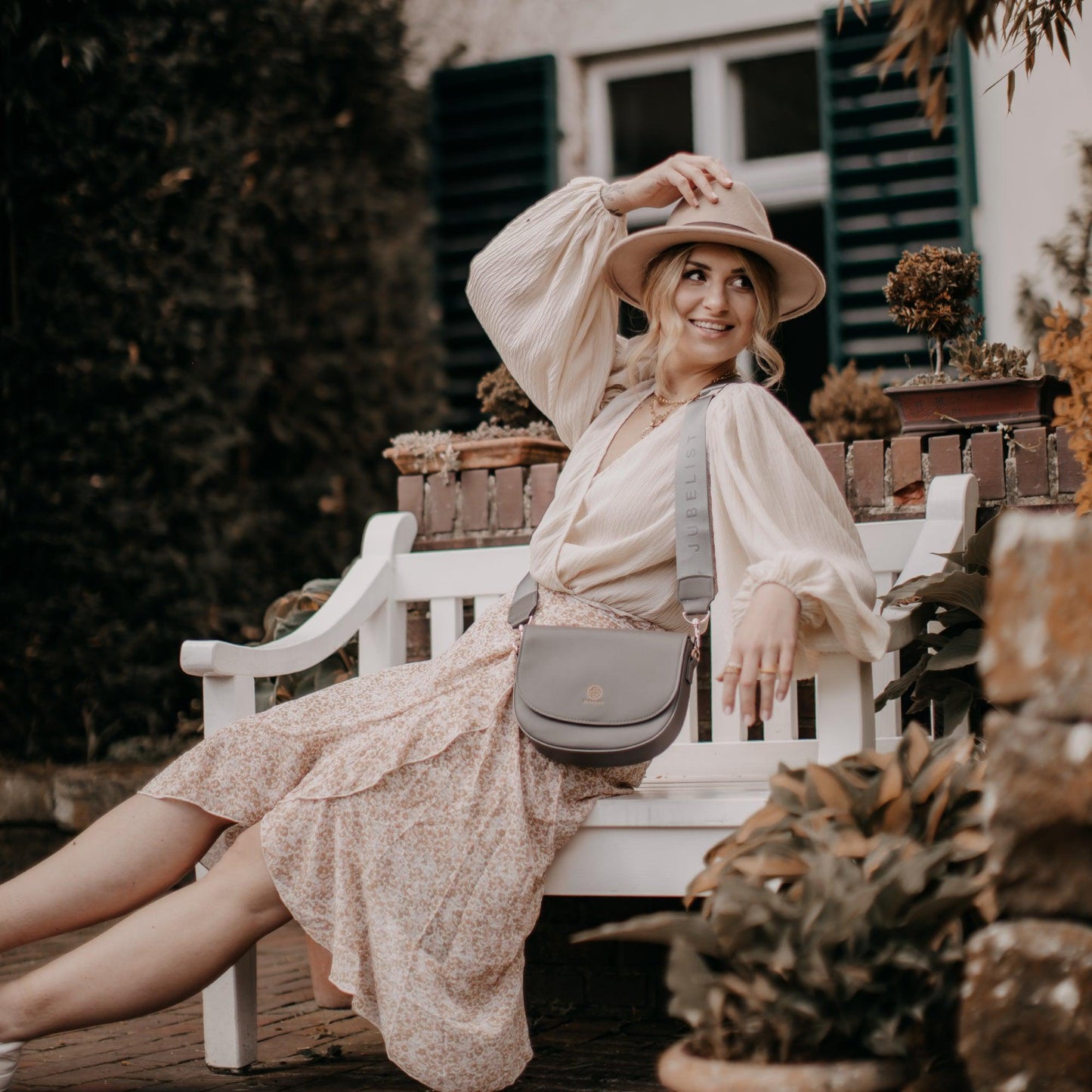 Eine junge Frau sitzt auf einer weißen Gartenbank in einem sommerlichen Outfit und trägt eine Saddle Bag in Graphite mit dem Fashion Strap in graphite