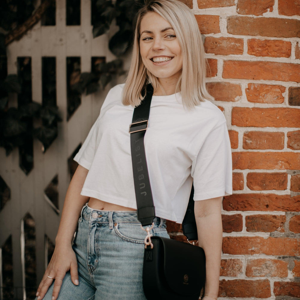 
                  
                    Eine junge Frau lehnt an einer Mauer und trägt den Fashion Strap schwarz an einer Saddle Bag in schwarz
                  
                