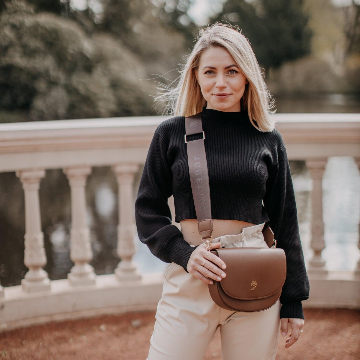 Eine junge Frau lächelt in die Kamera und trägt crossbody eine Saddle Bag in coffee mit einem passendem Fashion Strap in coffee