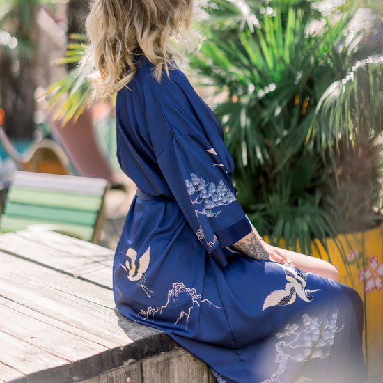 Ein Model sitzt in einen saphierblauen Kimono mit Kranichen und chinesischen Bäumen drauf vor einem exotischen Hintergrund
