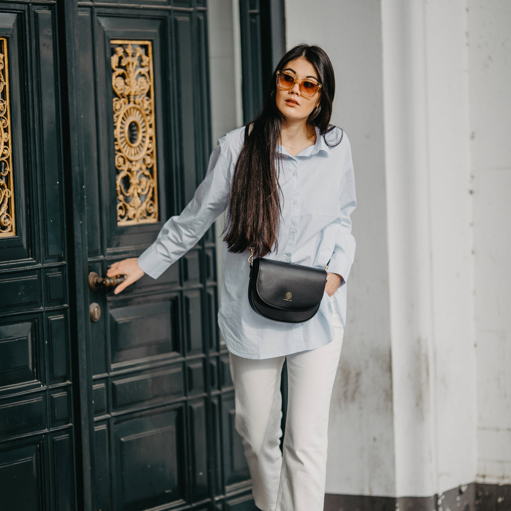 
                  
                    Eine junge Frau verlässt ein Haus durch eine imposante Tür und trägt die Saddle Bag in schwarz crossbody
                  
                